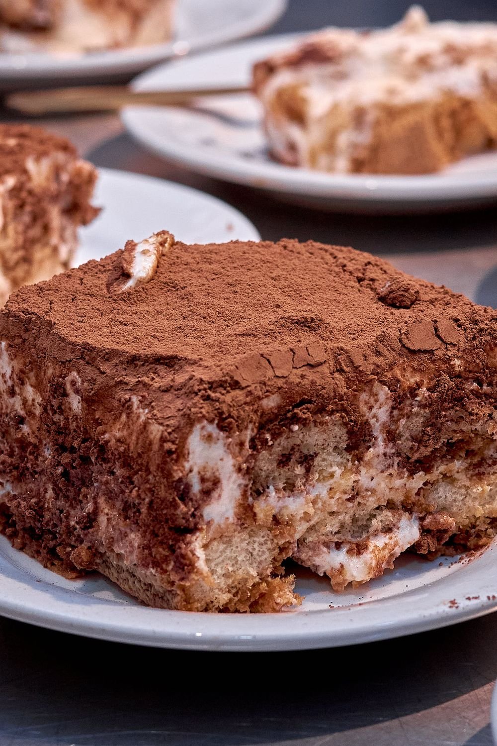 A cocoa-dusted slice of tiramisu with visible cream and sponge layers on a white plate, with more slices behind it.