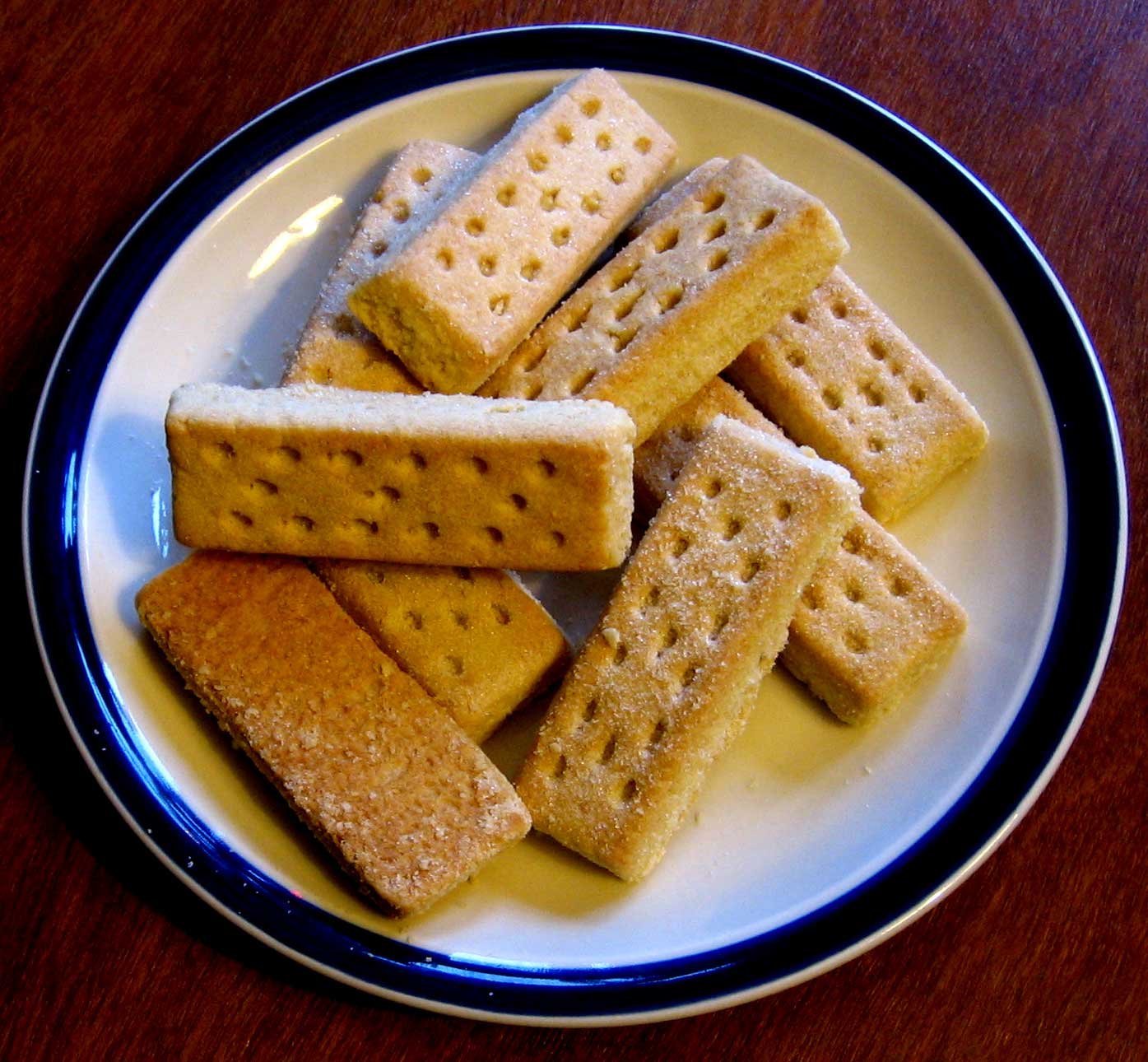 A plate of golden Scottish shortbread fingers dusted with sugar, pricked with fork holes, stacked on a blue-rimmed plate.