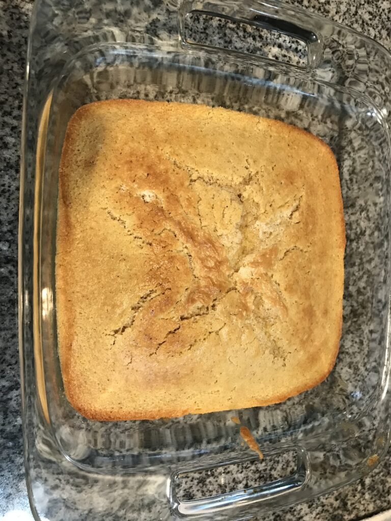 A golden baked cornbread casserole in a clear glass baking dish on a speckled countertop, showing its cracked golden top