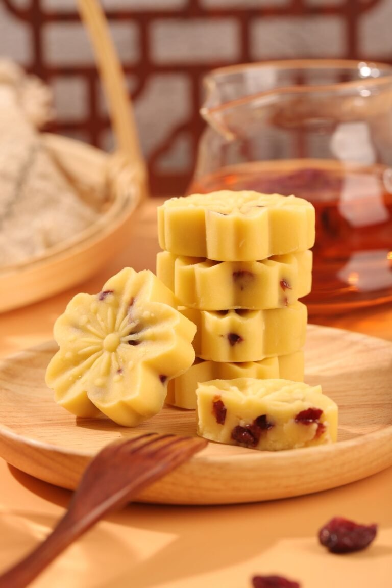 Flower-shaped pale shortbread biscuits studded with dried cranberry pieces, stacked on a wooden plate beside a teapot