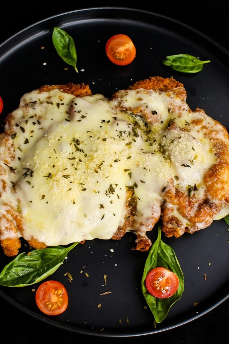 A plate of chicken parmesan topped with melted cheese and herbs, served with cherry tomatoes and fresh basil leaves.