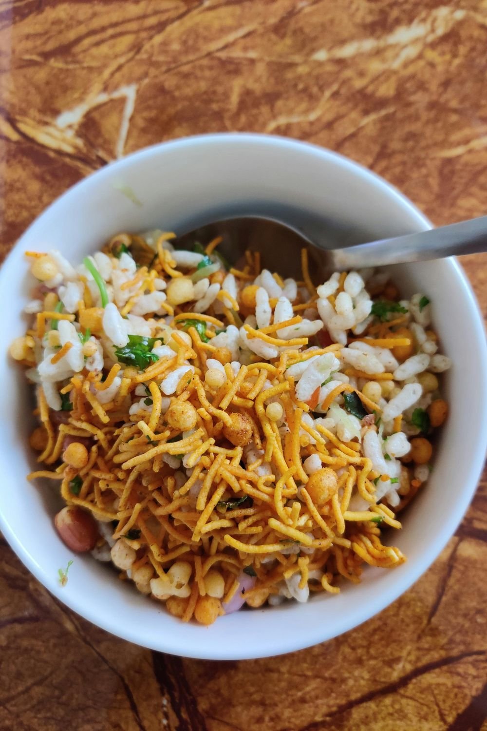 A white bowl of Indian snack mix with puffed rice, golden sev, peanuts, and herbs, with a spoon resting in the bowl.