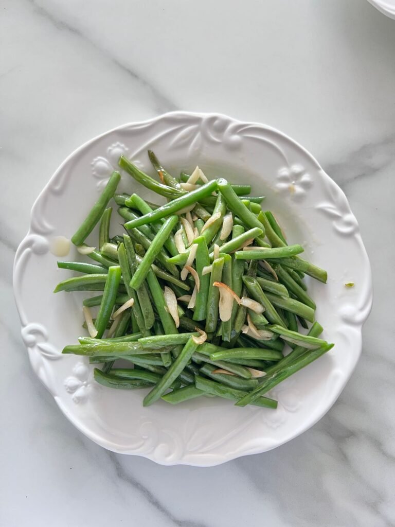 A white serving plate piled with glossy cooked green beans tossed with sliced garlic and toasted almonds on a marble table.