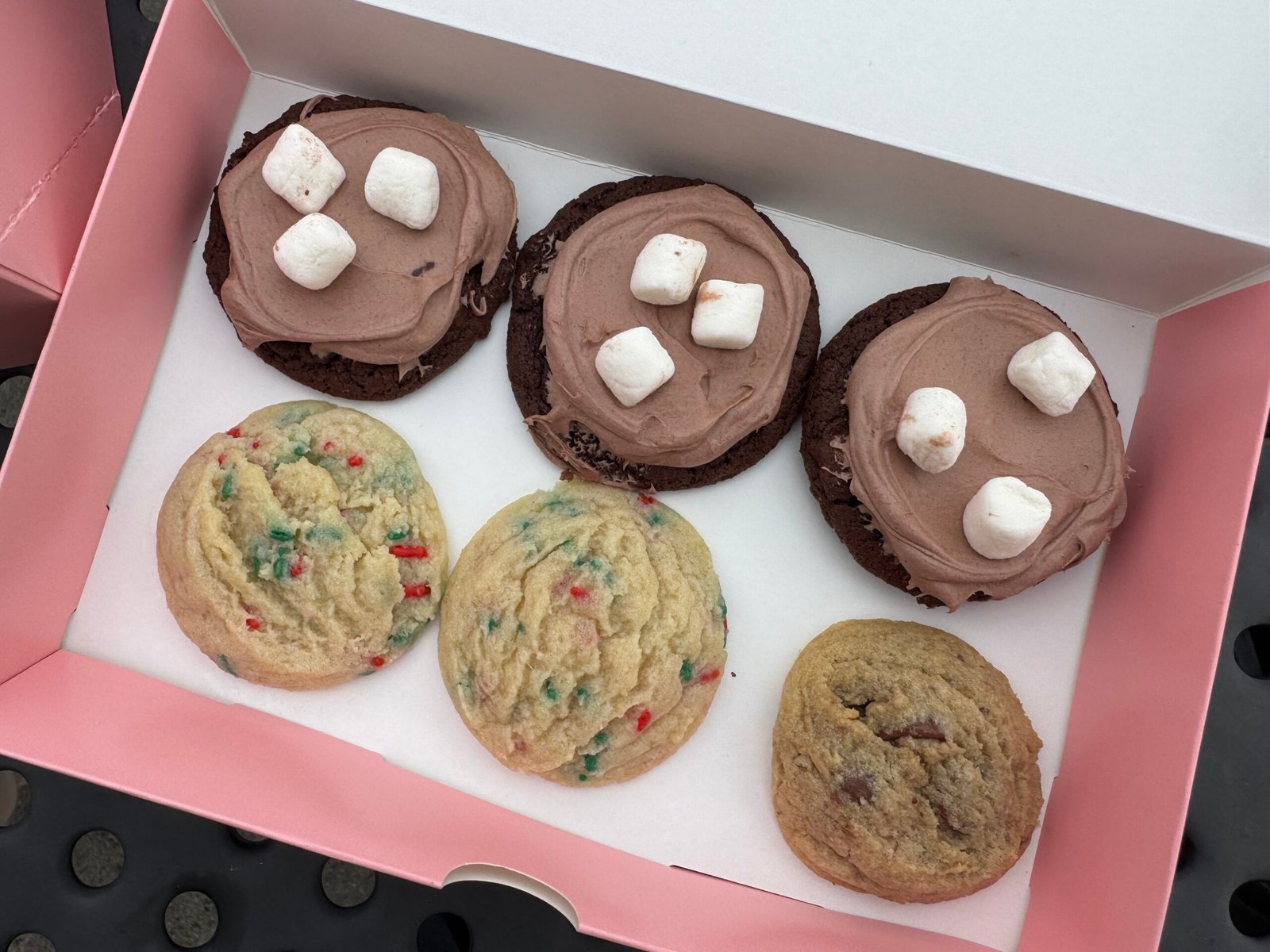 Hot Cocoa Cookies (Chocolate, Frosted, Topped With Marshmallows) 2 Dark chocolate cookies topped with chocolate frosting and mini marshmallows, in a pink bakery box with other cookies.