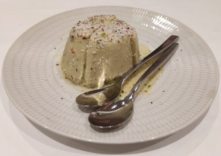 A moulded pistachio kulfi topped with chopped pistachios and coconut flakes on a white plate with two spoons