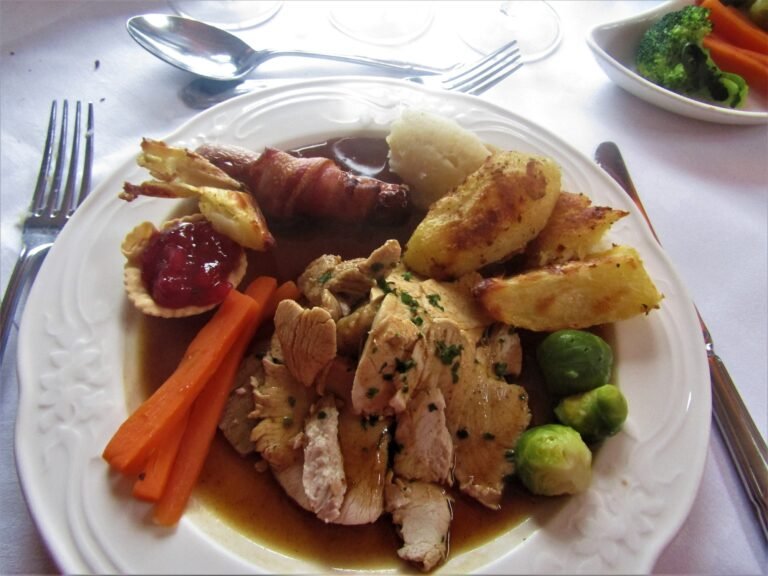 A plate of sliced roast turkey with brown gravy pooled around it, roast potatoes, carrots, and vegetables on the side