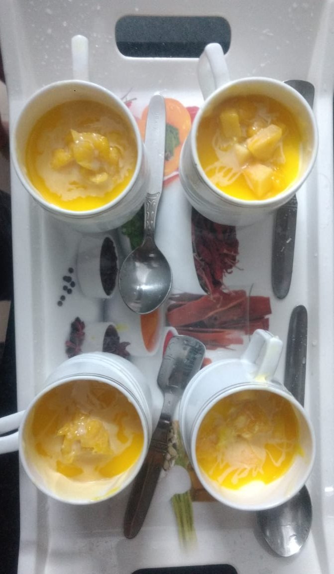 Two white cups of golden mango mousse topped with diced mango pieces, served on a printed tray with spoons