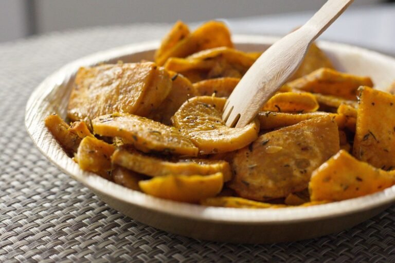 A close-up bowl of glossy roasted sweet potato slices glazed with maple and cinnamon, with a wooden serving fork resting in the bowl.