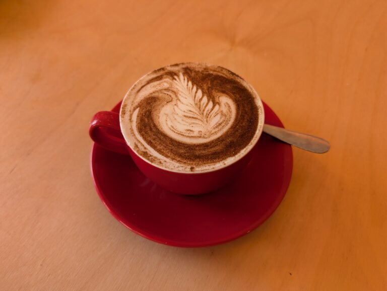 A red cup of chocolate chai with cocoa-dusted foam and a leaf pattern, served on a red saucer on a wooden table
