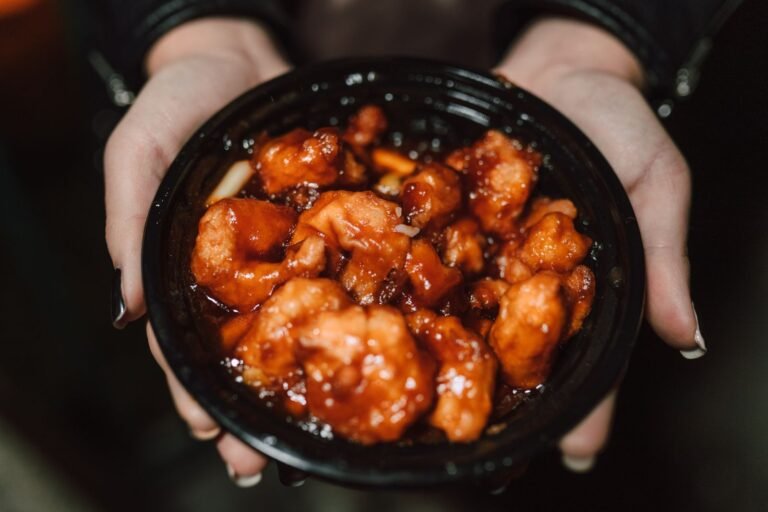 Hands holding a black bowl of glossy orange chicken pieces coated in a sticky, sweet orange glaze, ready to serve over rice