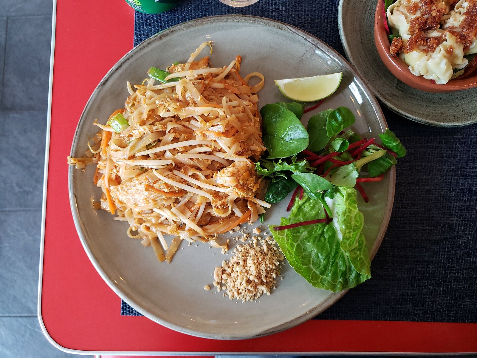 Pad Thai (Better Than Takeout in 30 Minutes) 2 A gray plate of pad thai rice noodles with bean sprouts, crushed peanuts, a lime wedge, and fresh greens on a red table