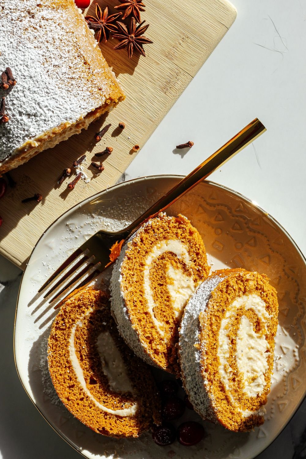Pumpkin Roll 2 Slices of pumpkin roll with a cream cheese swirl on a plate, with the whole dusted pumpkin roll on a wooden board behind them.