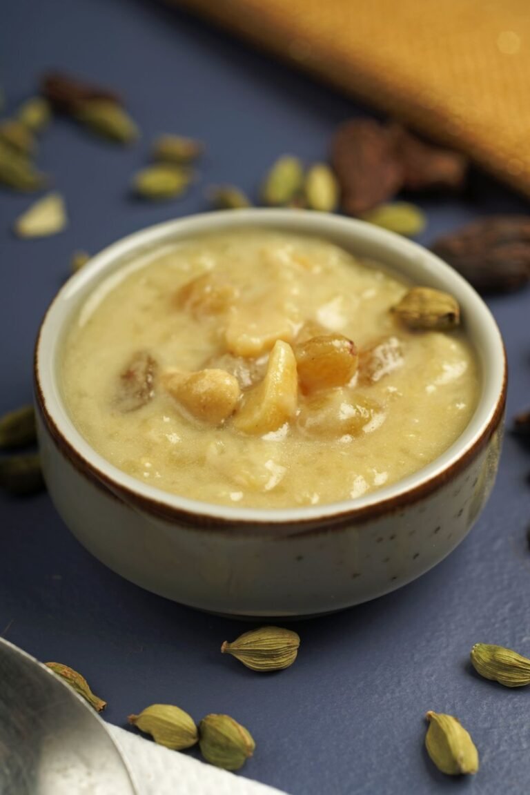 A ceramic bowl of golden saffron rice pudding topped with cashews, raisins, and cardamom pods on a dark blue surface.