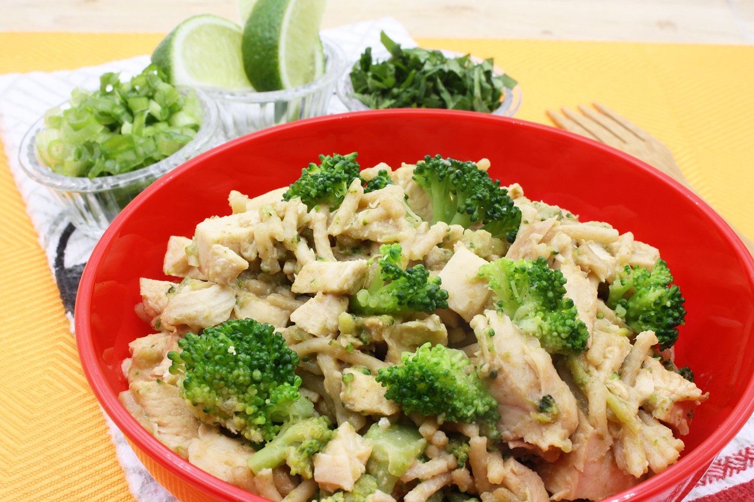 Thai Peanut Chili Noodles (30-Minute, Saucy, Boldly Spiced) 3 Peanut-sauce noodles with chicken and broccoli in a red bowl, with lime wedges and herbs in the background.
