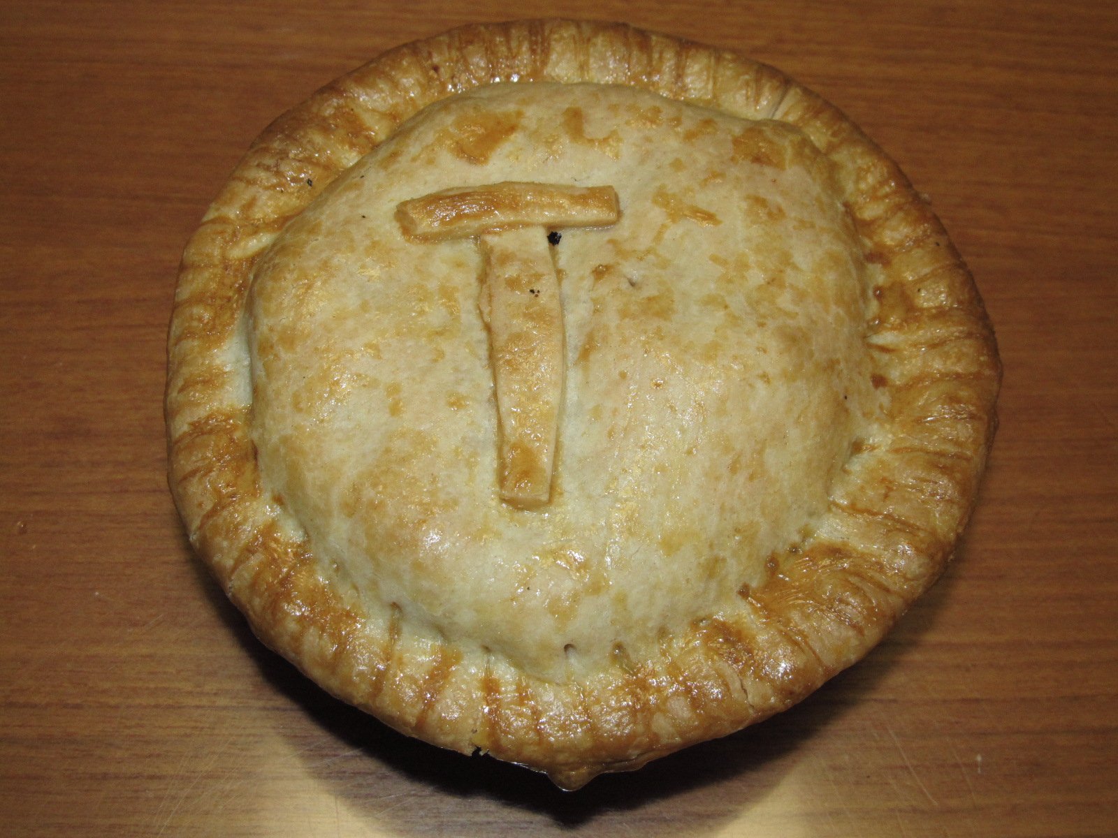 Turkey Pot Pie 2 A whole golden-brown turkey pot pie with a pastry letter T on its flaky crust, sitting on a wooden table