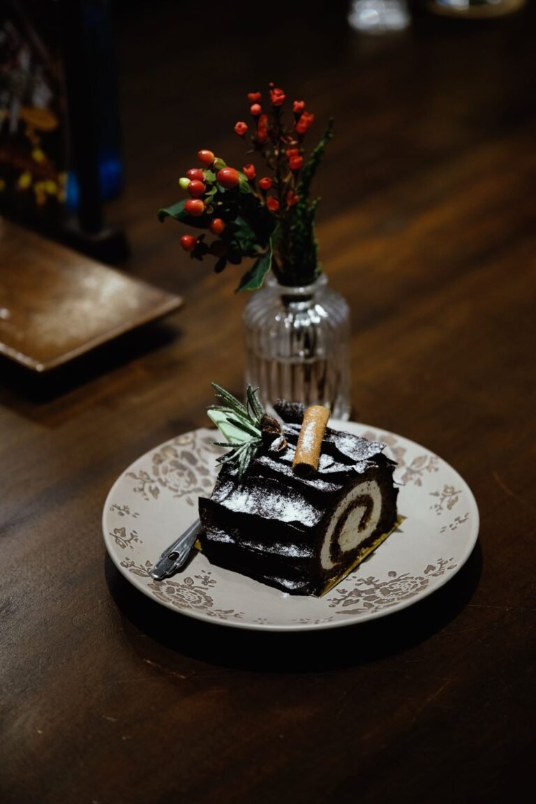 A chocolate yule log slice with a cream spiral, dusted with powdered sugar and topped with a cinnamon stick, on a patterned plate.